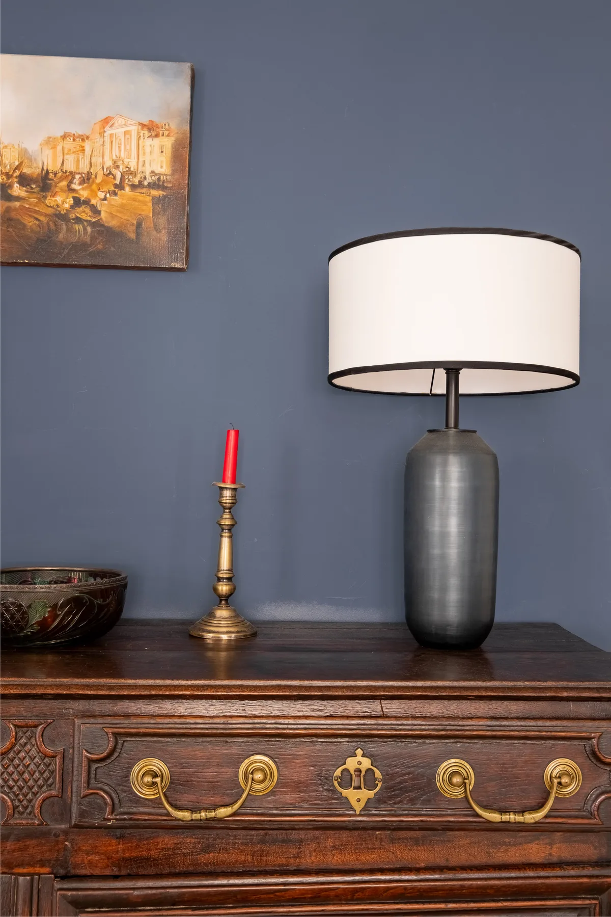 A table lamp and candle sit on a carved wooden dresser beneath a painting.