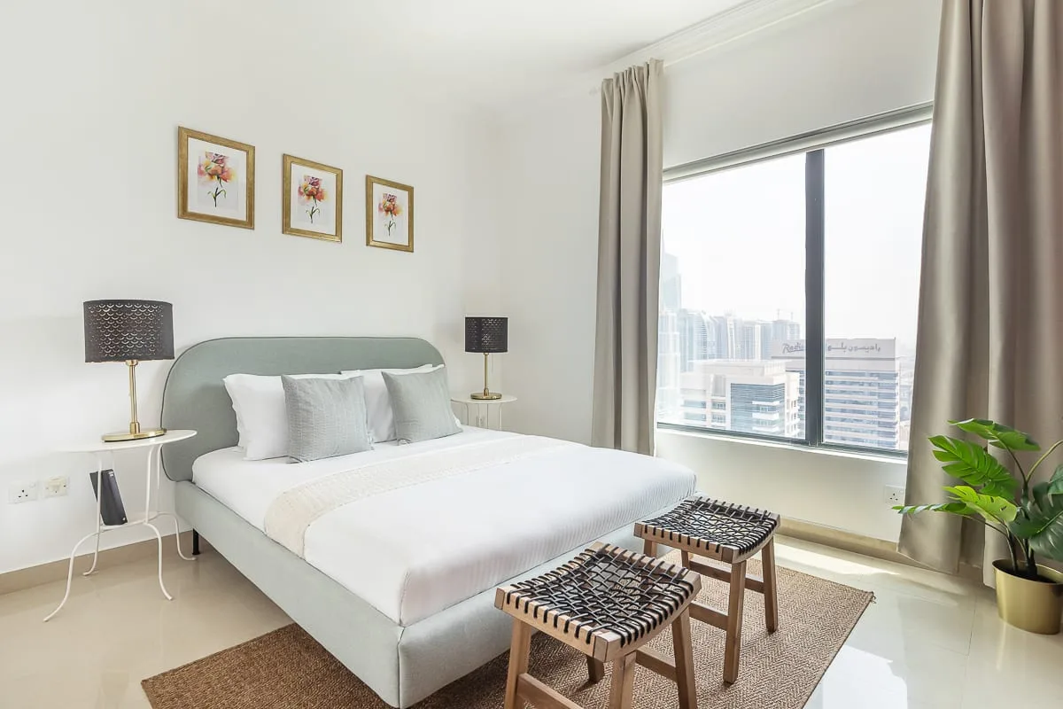 Un dormitorio luminoso con una cama bien tendida junto a una ventana grande.