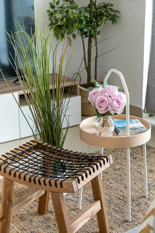 Una zona de estar con un taburete de madera tejido, plantas y flores sobre una mesa auxiliar.