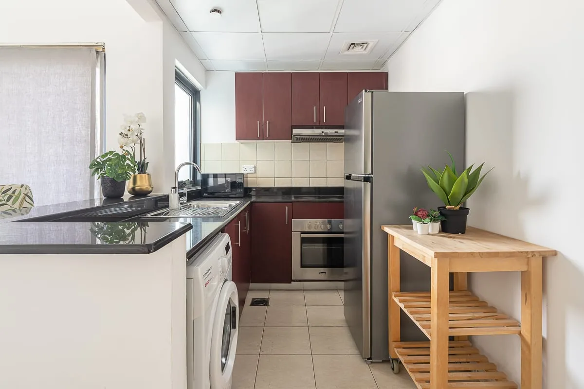 Una cocina compacta con gabinetes oscuros, un refrigerador y una mesa de estantes de madera.