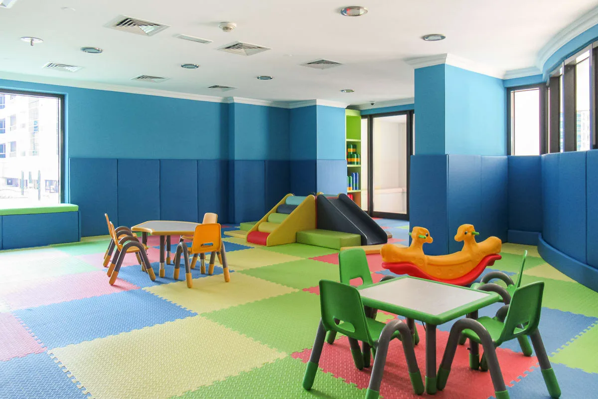 Una sala de juegos interior colorida tiene piso acolchado, mesas pequeñas y juegos infantiles.
