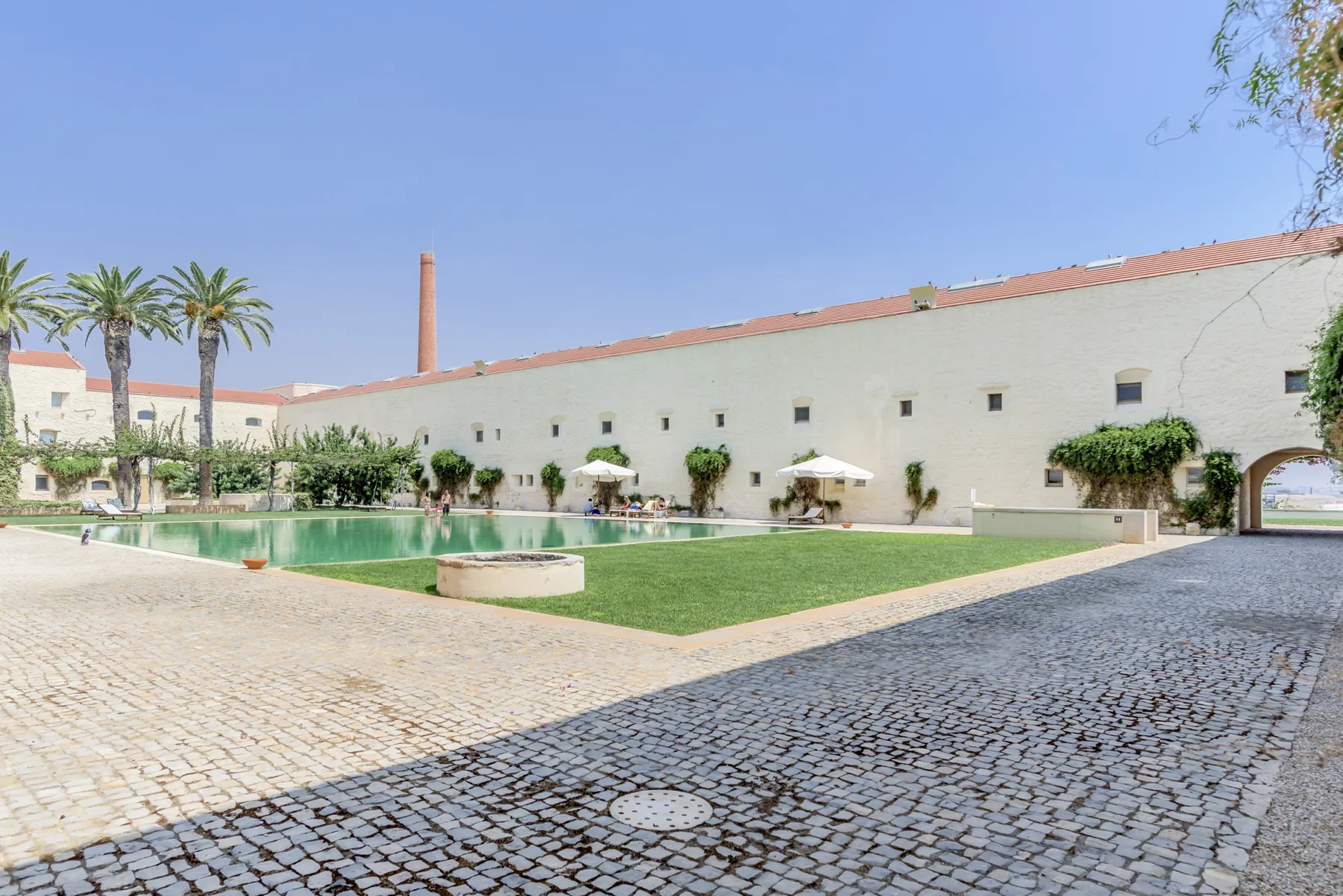 A swimming pool and lawn sit beside a long white building and cobbled courtyard.