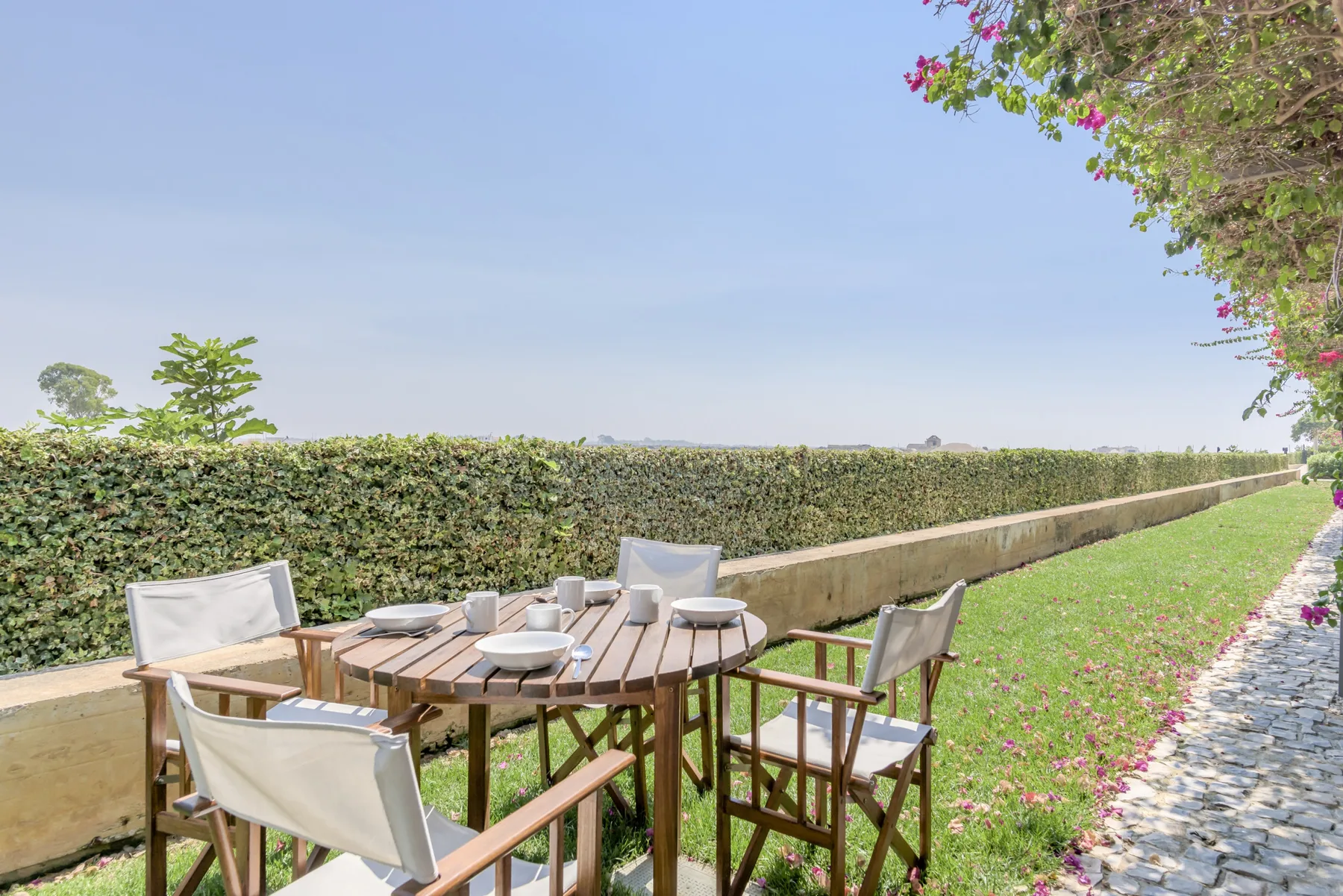 An outdoor dining area with wooden seating beside a hedge and grassy path.