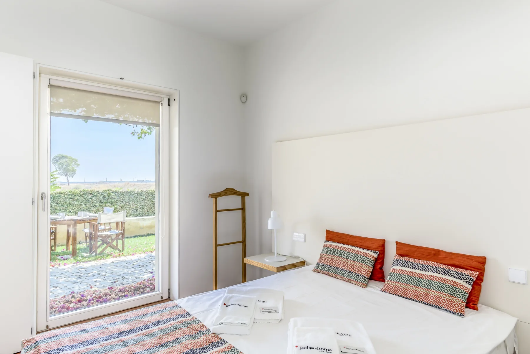 A bright bedroom with a bed beside a glass door opening onto a garden.
