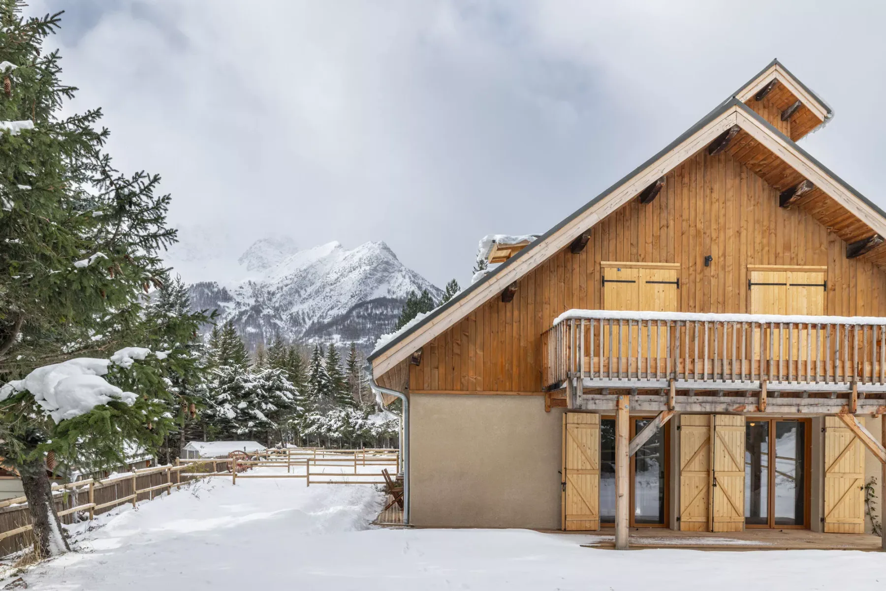 A wooden chalet stands beside a snow-covered yard, with mountains in the background.