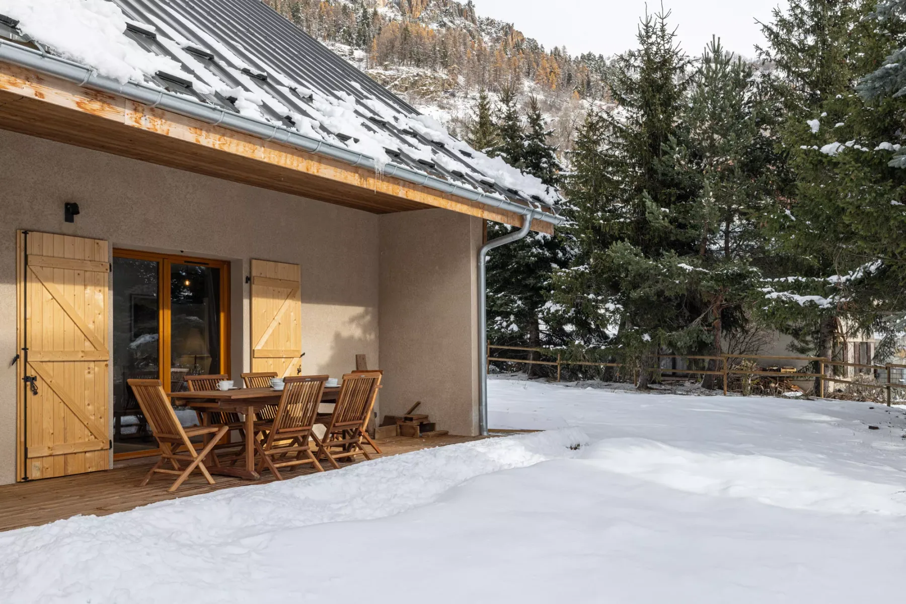 A snow-covered outdoor dining area beside a building, with a wooden table and seating.