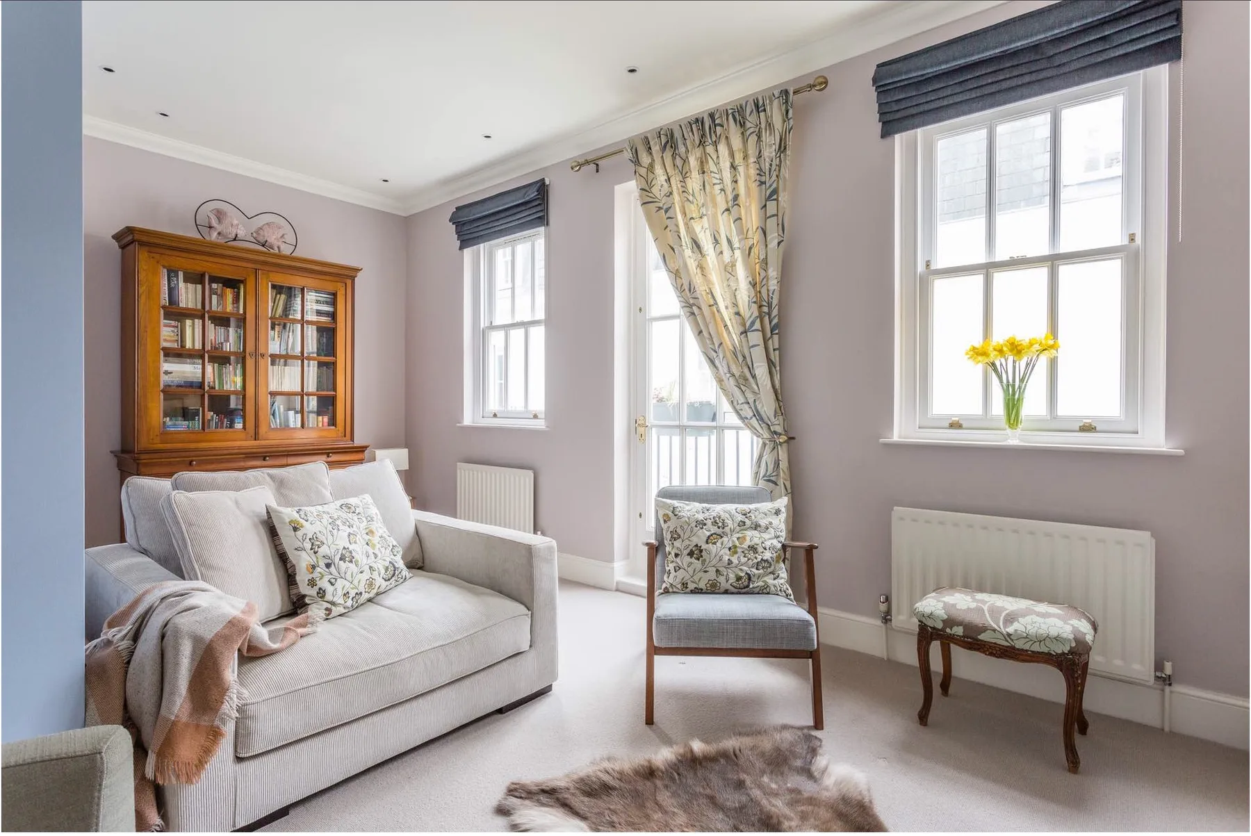 A bright living room with light seating, a wooden bookcase, and tall windows.