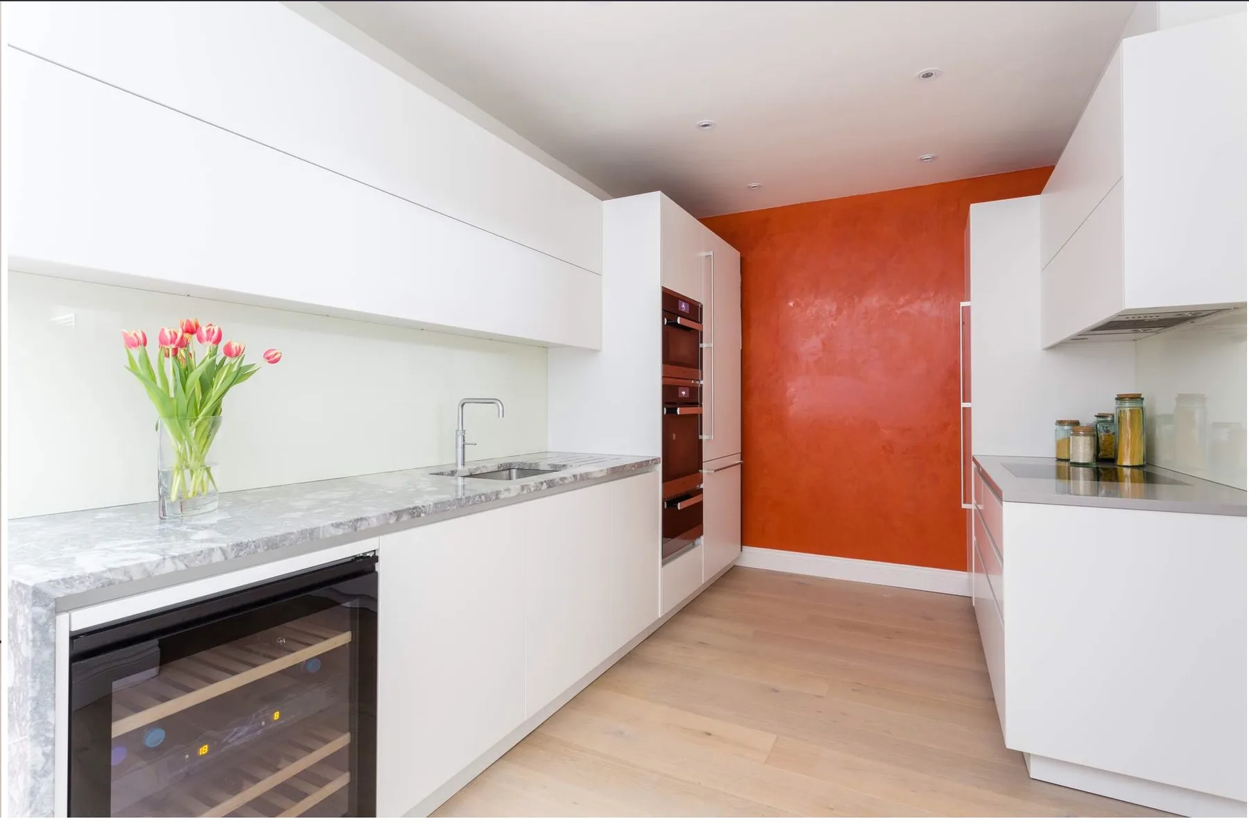 A modern kitchen with white cabinetry, a long counter, and a red accent wall.