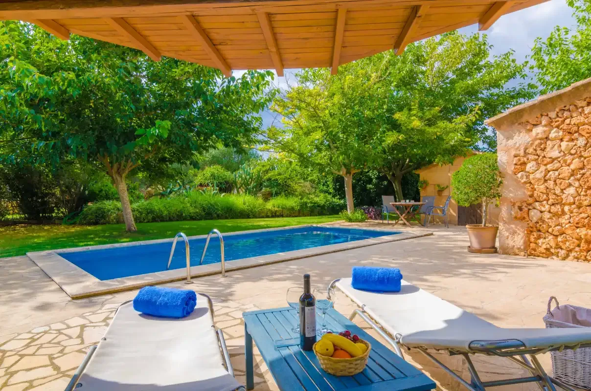 A swimming pool and loungers sit beneath a wooden pergola, surrounded by trees.