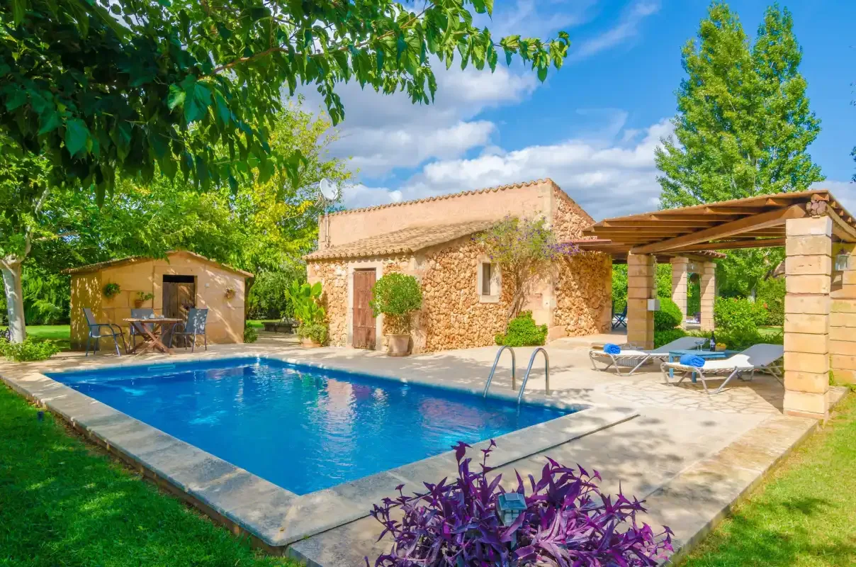 A swimming pool sits beside a stone house and shaded outdoor seating.