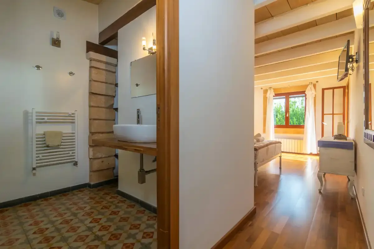 A bathroom with a sink opens onto a bright room with exposed ceiling beams.