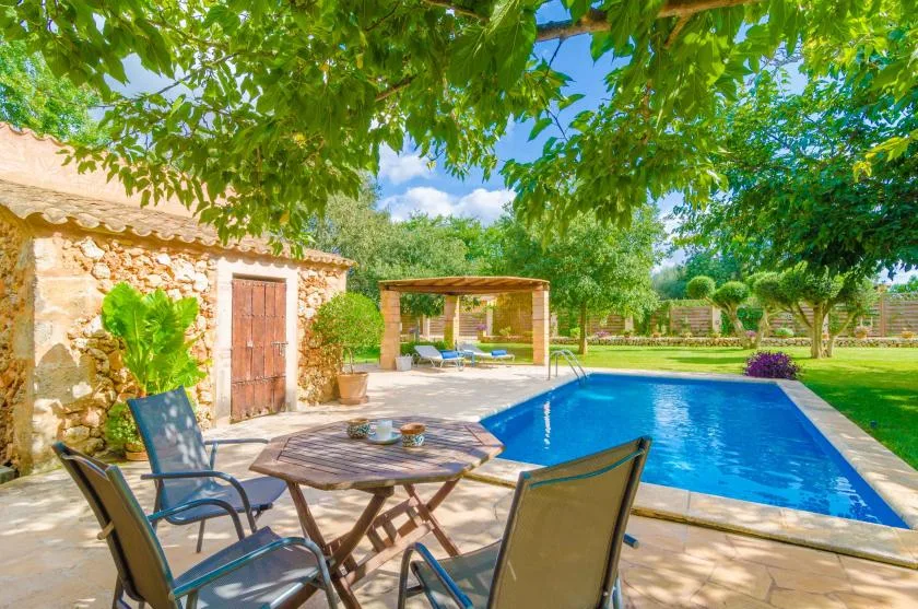 An outdoor pool beside a stone building, with shaded seating beneath leafy trees.