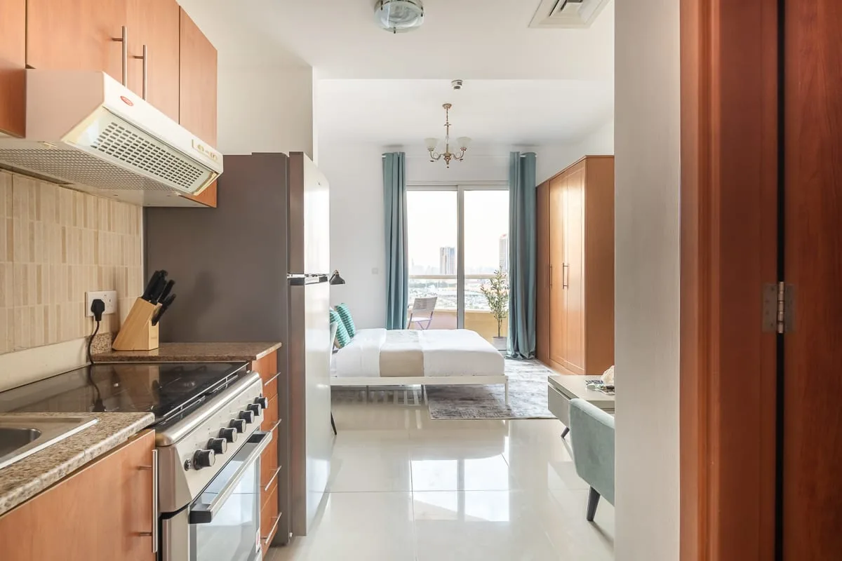 A kitchen opens onto a bedroom with a bed and large windows.
