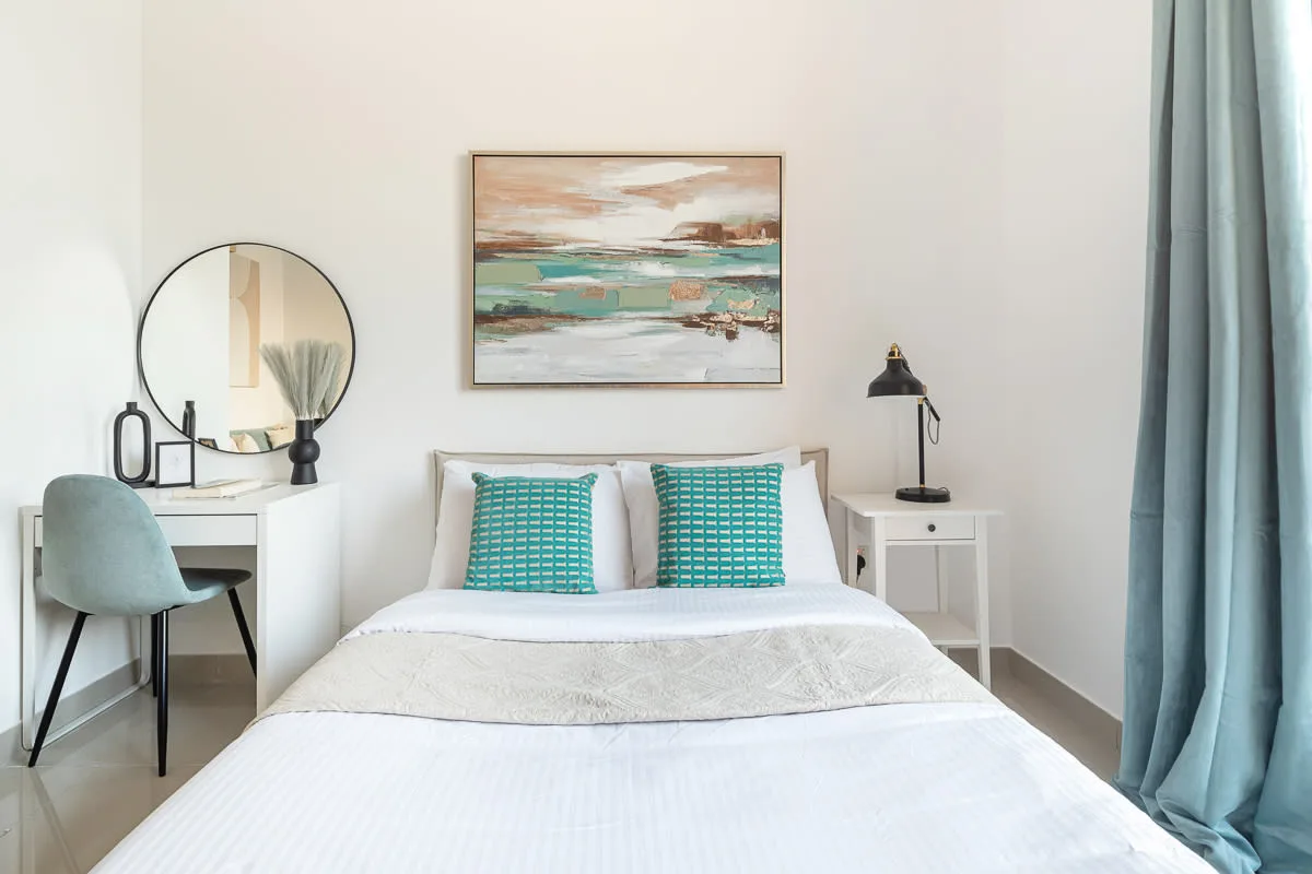 A bright bedroom with a bed, turquoise pillows, and artwork above the headboard.