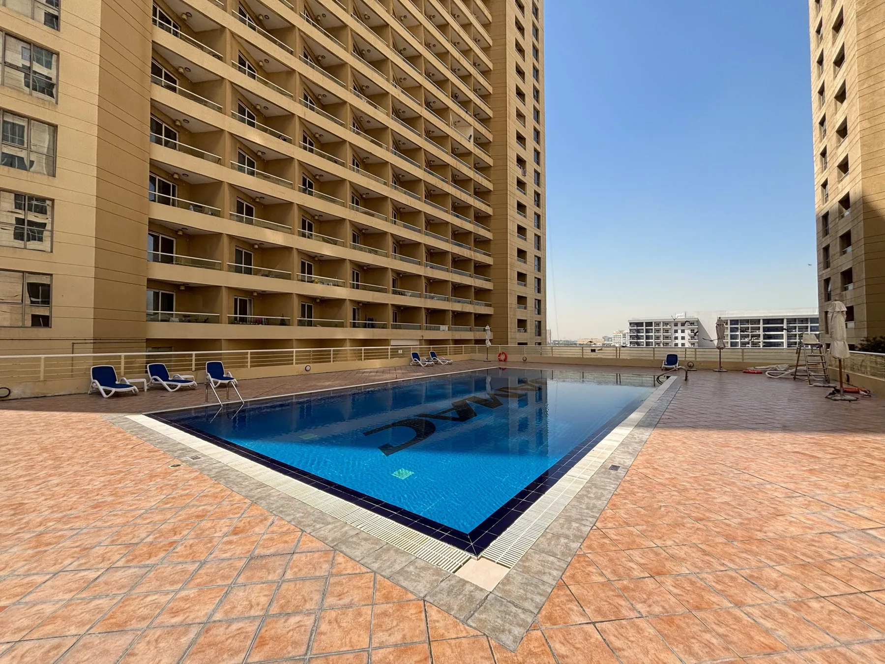 An outdoor swimming pool sits between tall apartment buildings on a tiled terrace.