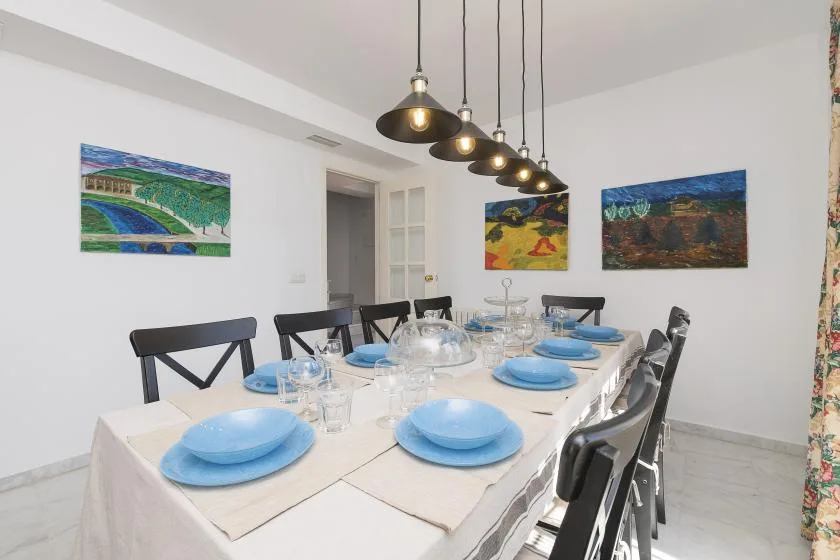 A dining room has a long table set with blue plates beneath pendant lights.