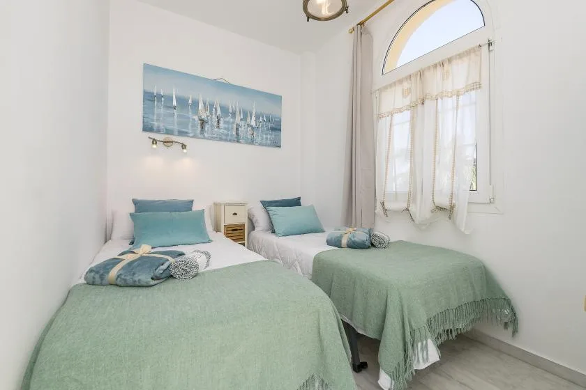 A bright bedroom with two beds dressed in pale green bedding beside a curtained window.