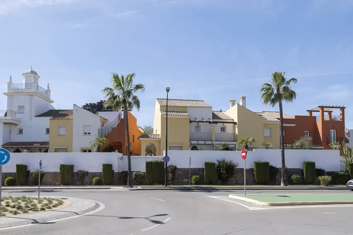 A residential street lined with colorful houses, palm trees, and low hedges.