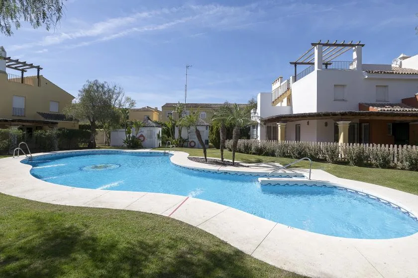 A curved outdoor swimming pool sits beside a lawn and low-rise buildings.