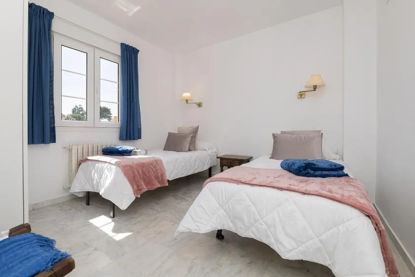 A bright bedroom with two single beds, blue curtains, and white bedding.