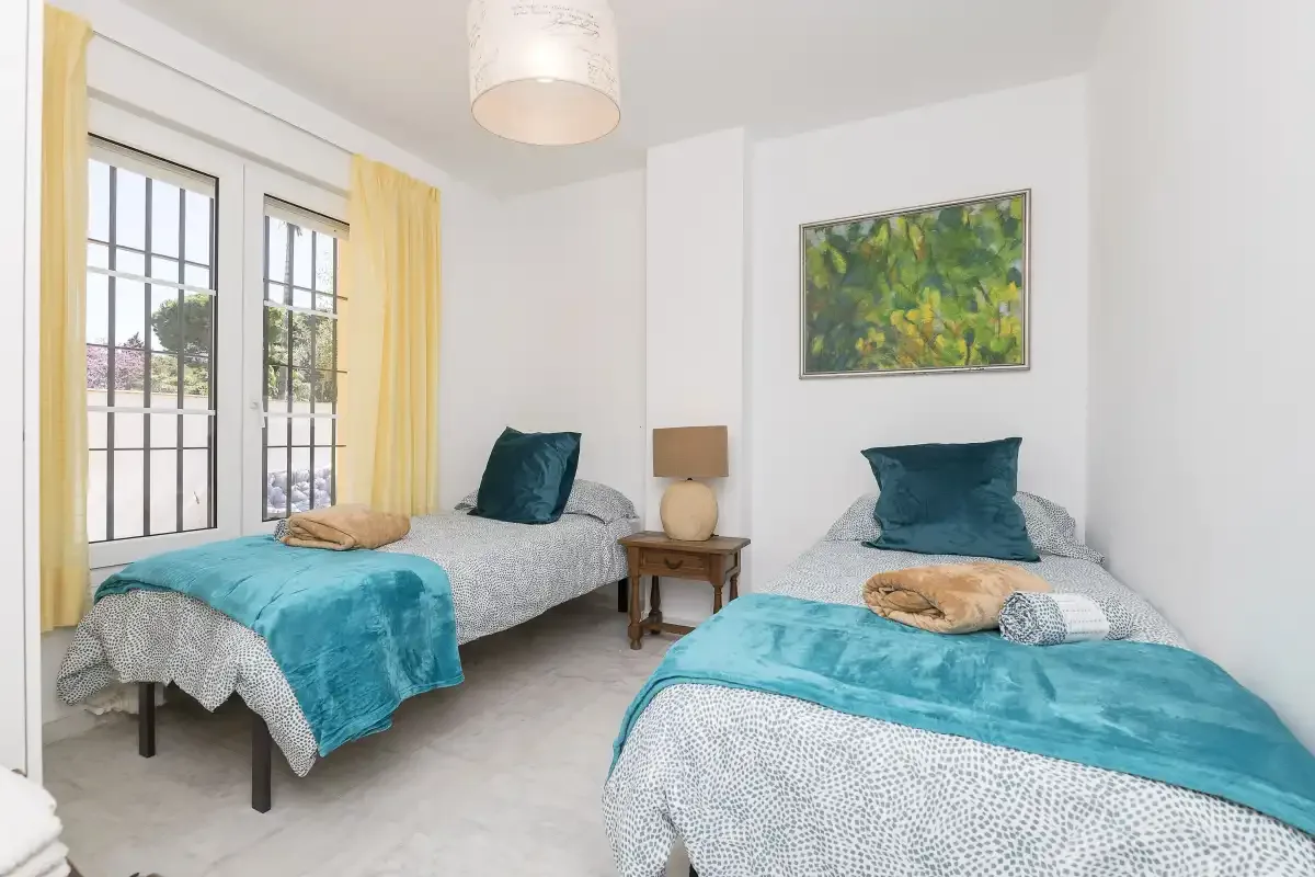 A twin bedroom with two beds, teal bedding, and windows with yellow curtains.