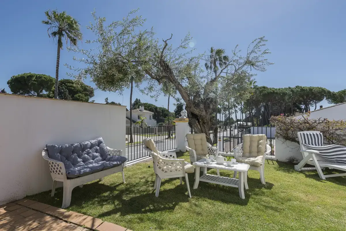 A garden seating area with white chairs and a table beneath leafy trees.