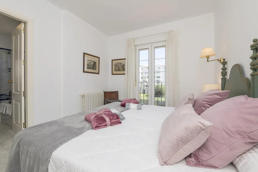 A bedroom with a bed dressed in white and pink linens beside tall glass doors.