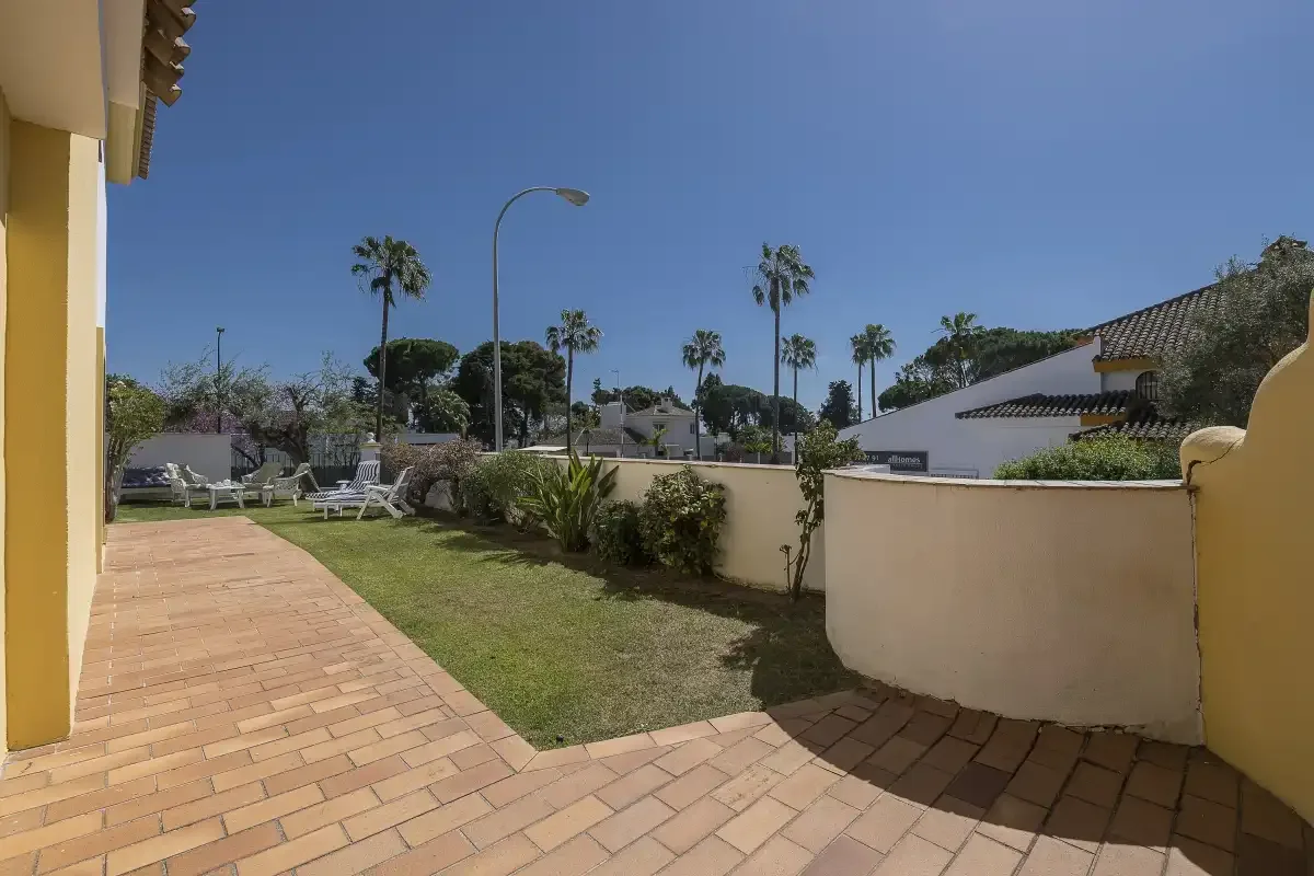 A paved patio borders a small lawn, with trees and buildings beyond.