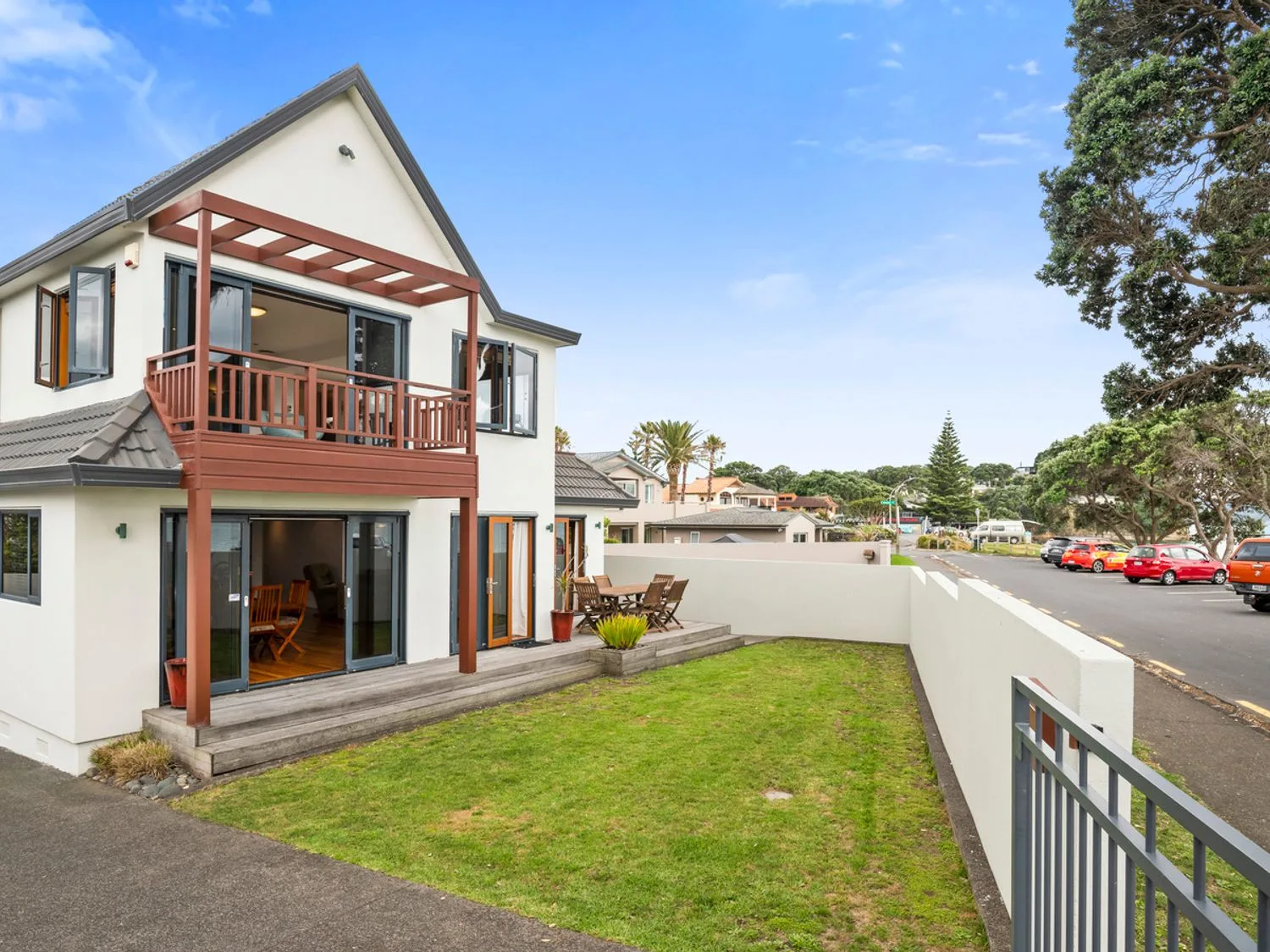 Una casa de dos plantas con un pequeño balcón y una zona de patio con vistas a un jardín con césped y a una calle con coches aparcados en Remarkable Red Beach - Red Beach Holiday Home, en Whangaparaoa.
