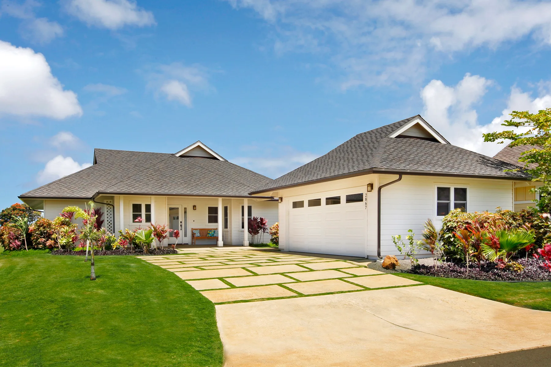 A single-story house with a driveway bordered by lawn and flowering plants.