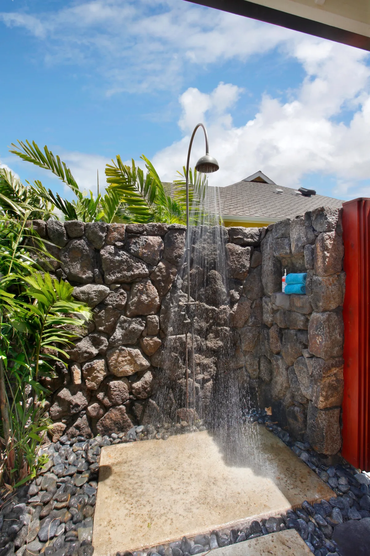 An outdoor shower stands against a stone wall beside tropical plants.