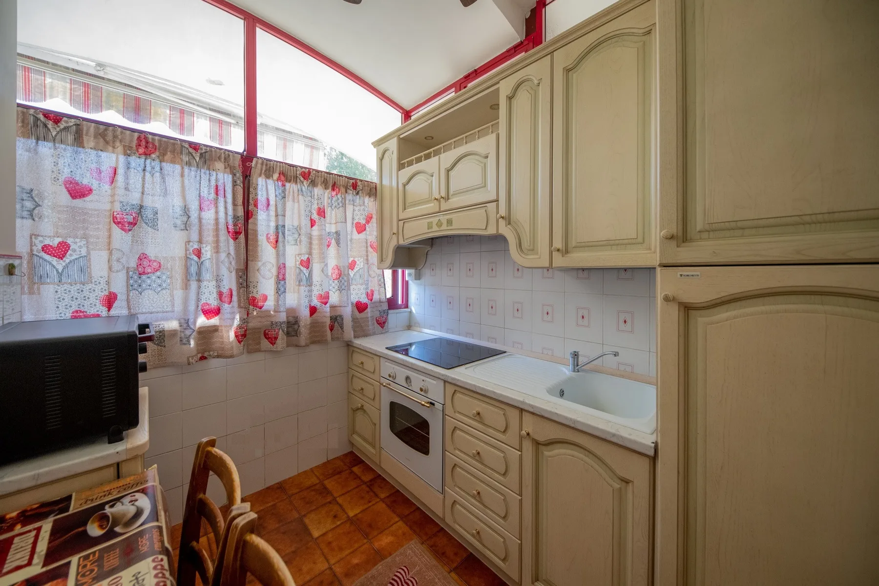 A compact kitchen with cream cabinetry, a cooktop, and a sink beneath curtained windows.