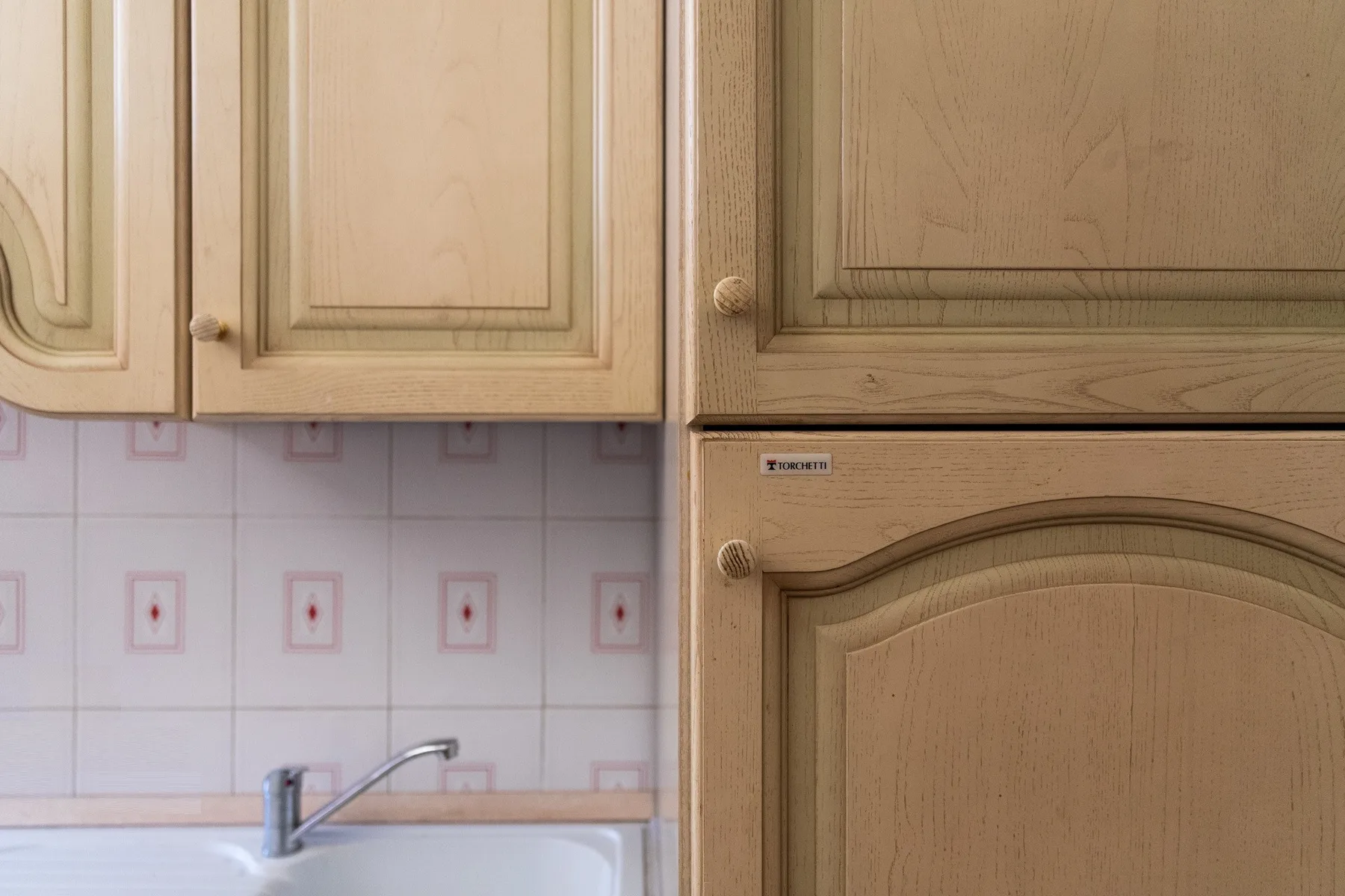 A kitchen sink sits beneath wooden cabinets, with patterned wall tiles behind it.