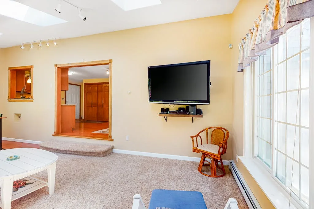 A bright living room with a wall-mounted television and large windows.