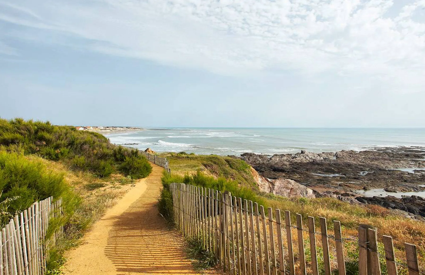 A sandy coastal path runs between low vegetation and fencing beside the rocky shore.
