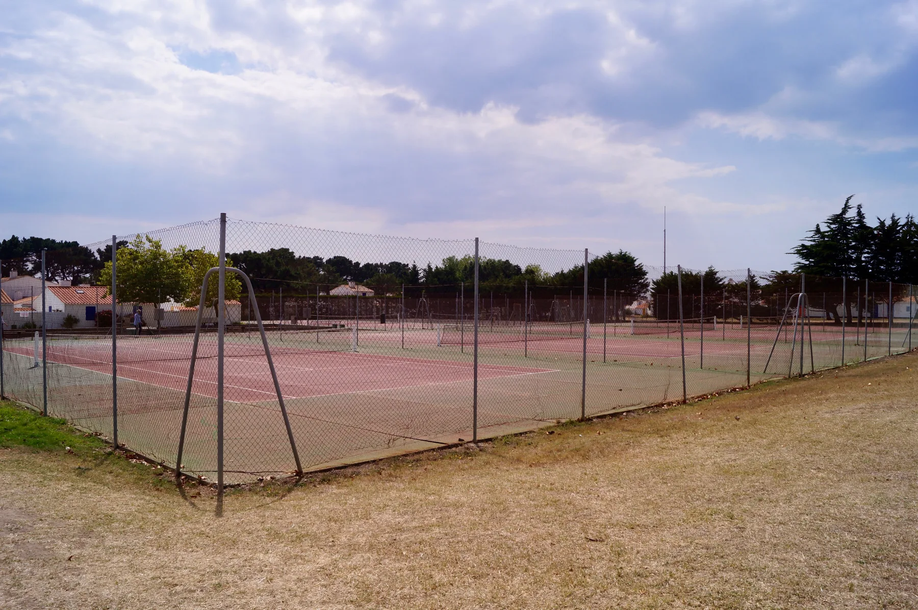 An outdoor tennis court sits behind a chain-link fence beneath a cloudy sky.