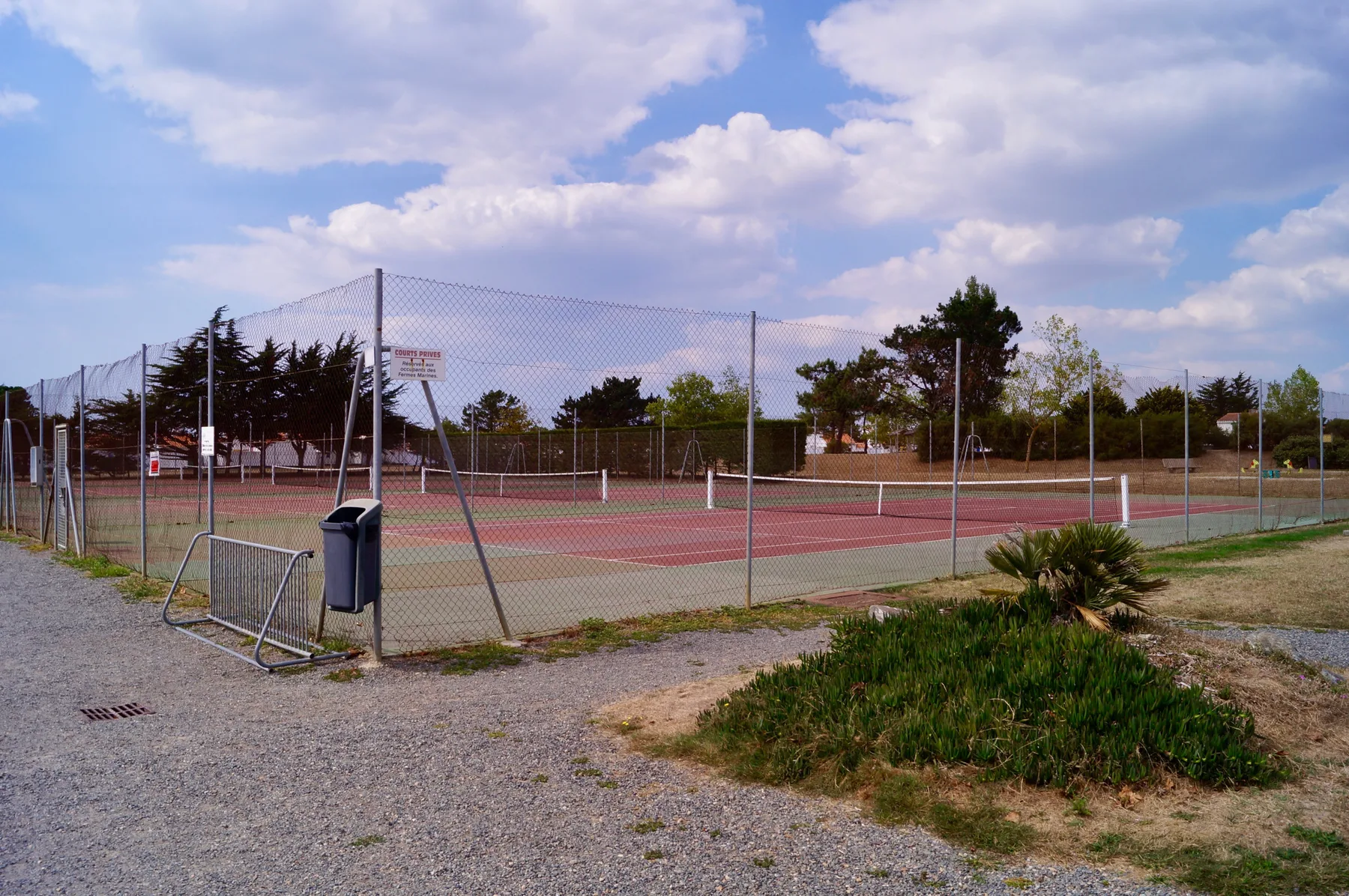 A fenced outdoor tennis court sits beside a gravel path and grassy area.