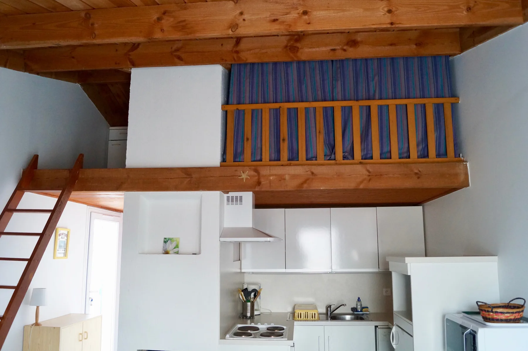 A compact kitchen sits beneath a wooden loft with a ladder and railing.