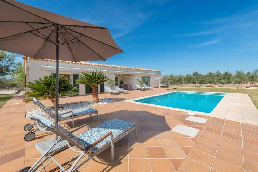 Una piscina al aire libre junto a una casa baja, con tumbonas en el patio embaldosado.
