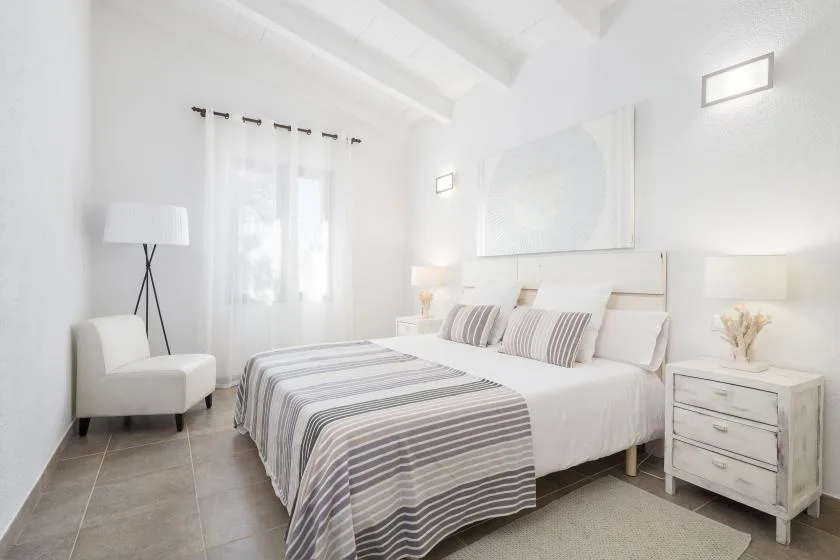 Un dormitorio luminoso con una colcha a rayas y un sillón blanco junto a la ventana.