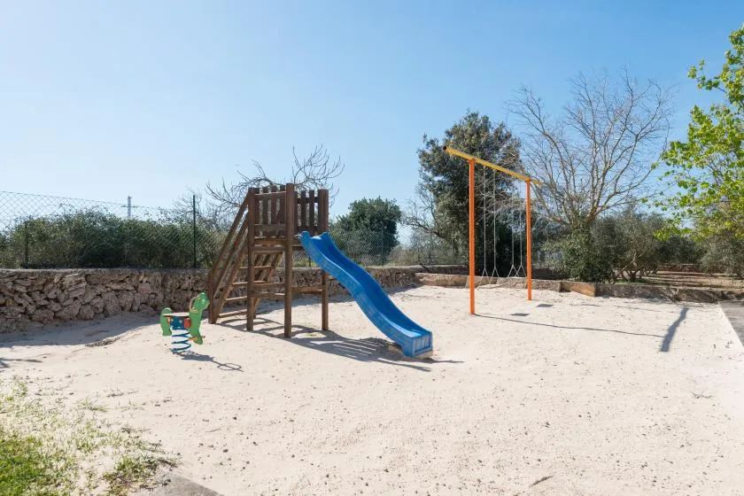 Un parque infantil al aire libre con estructura de madera, tobogán azul y columpios sobre arena.