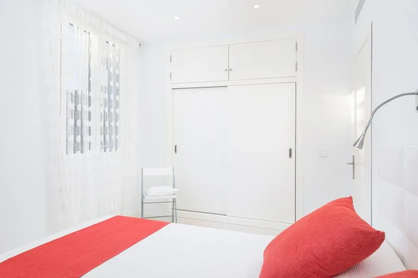 Un dormitorio luminoso con una cama, ropa de cama roja y cortinas translúcidas.