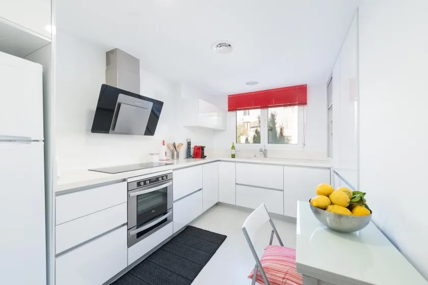 Una cocina blanca y luminosa con una ventana y una persiana roja sobre el fregadero.