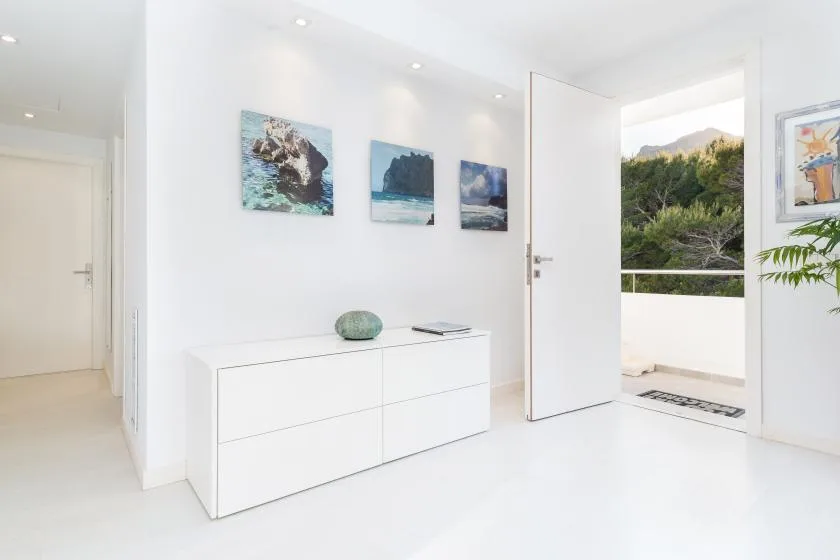Un pasillo blanco y luminoso con un mueble bajo y una puerta abierta hacia la vegetación.