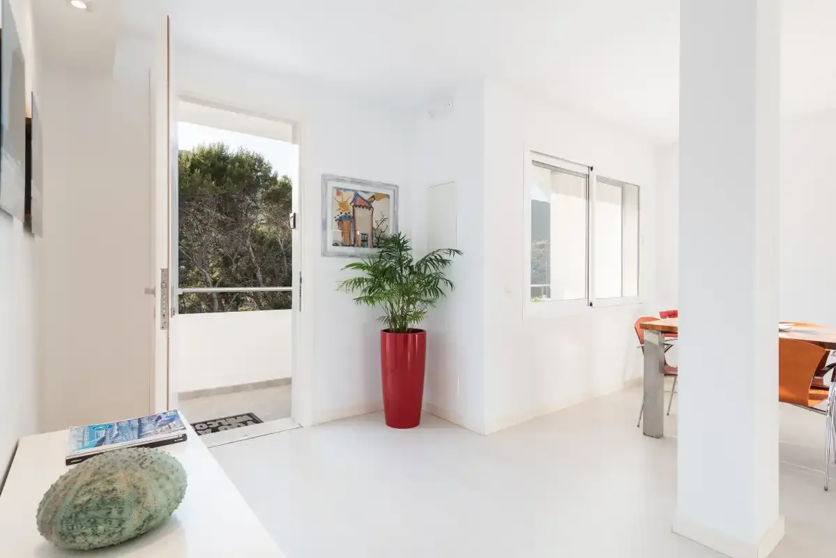 Un interior blanco y luminoso se abre a un balcón, con una planta junto a la puerta.
