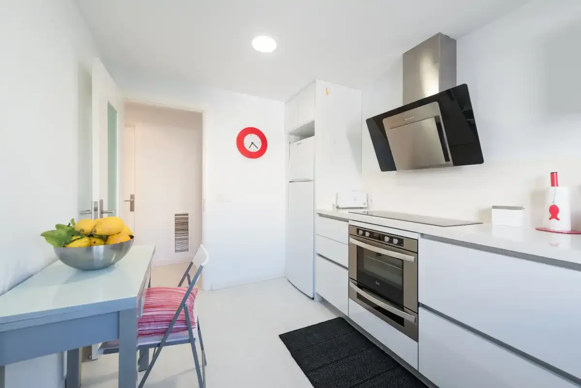 Una cocina blanca y luminosa con horno, refrigerador y una pequeña mesa de comedor.