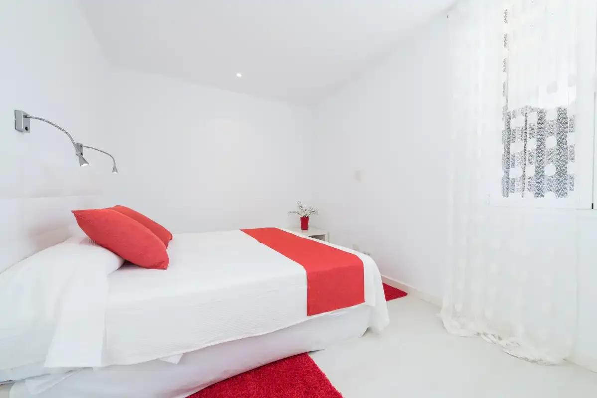 Un dormitorio luminoso con una cama blanca y detalles rojos.