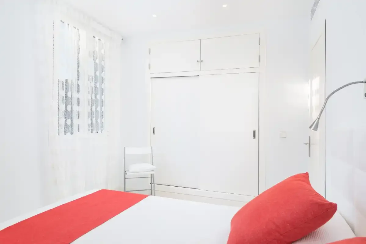 Un dormitorio luminoso con ropa de cama blanca y almohadas rojas junto a una ventana con cortinas.