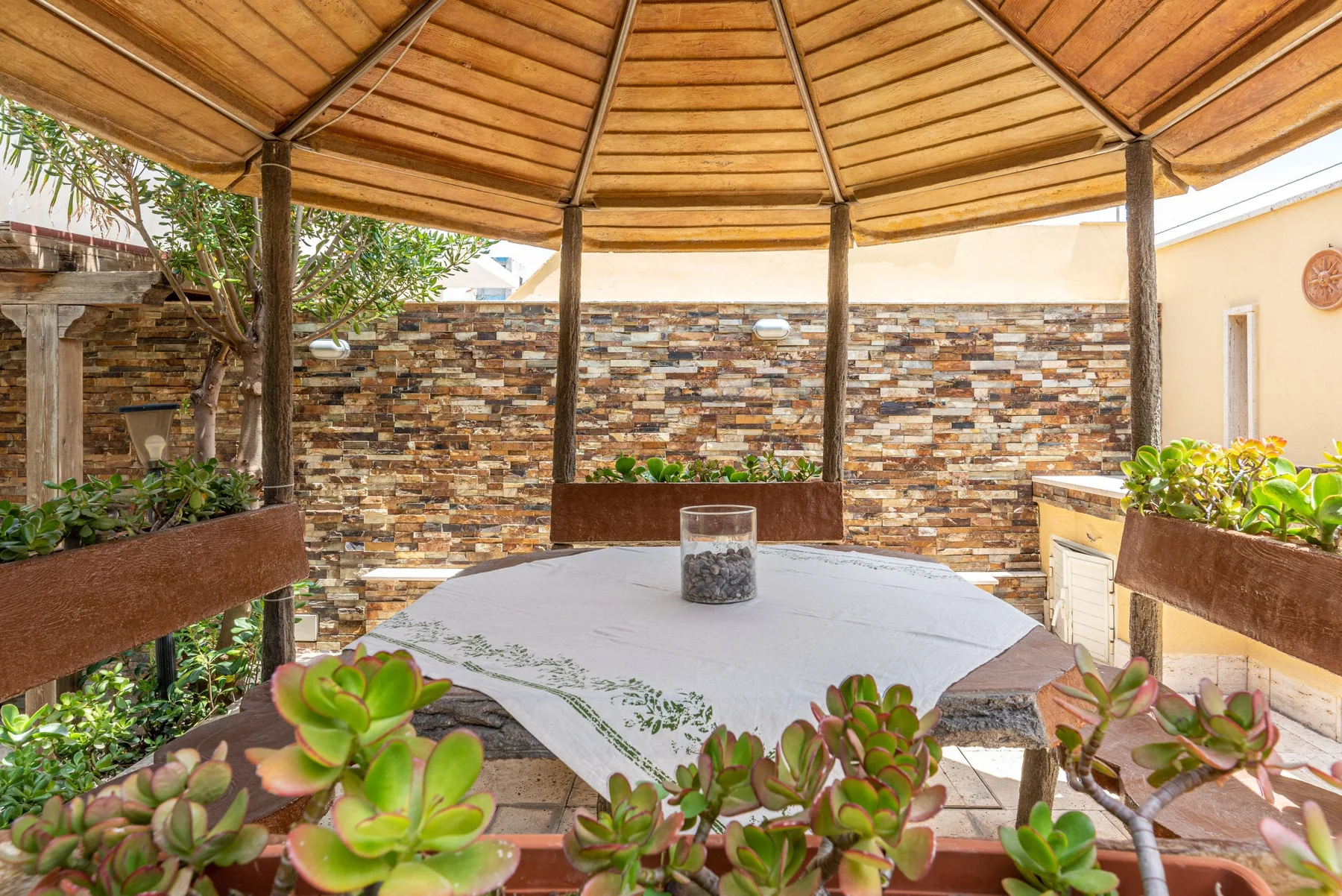 Una zona de estar al aire libre está bajo una pérgola de madera, junto a un muro con patrón de piedra.