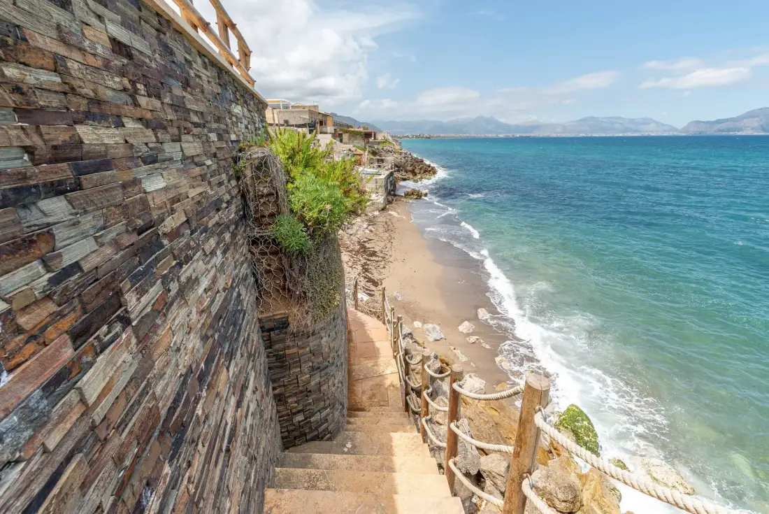 Unas escaleras de piedra bajan junto a un muro costero hasta una playa de arena y aguas turquesas.